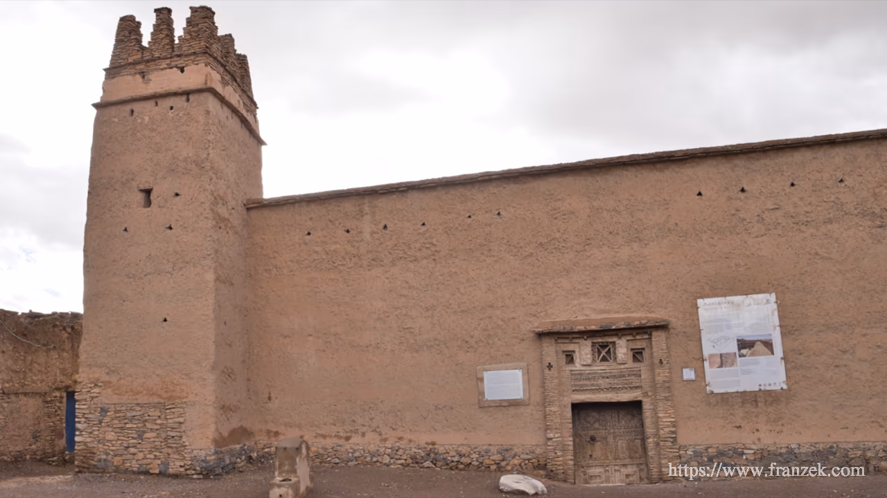 Süd-Marokko Mietwagen-Route: Kosten & Tipps (Anti-Atlas) 32 Außenansicht des Agadir von Ait Kine unter bewölktem Himmel. Das massive, fensterlose Gebäude besteht aus rötlichem Stampflehm auf einem Steinsockel. Links ragt ein quadratischer Wehrturm mit Zinnen auf. Rechts befindet sich ein kleinerer Eingang mit verzierter Holztür und geometrischen Ornamenten im Mauerwerk darüber. An der Fassade ist eine weiße Informationstafel angebracht.