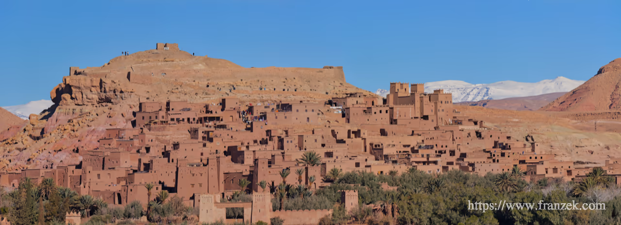 Süd-Marokko Mietwagen-Route: Kosten & Tipps (Anti-Atlas) 61 Weitwinkelaufnahme des Ksar Ait Ben Haddou. Ein rötlich-brauner Hügel, der dicht mit traditionellen Lehmbauten (Kasbahs) bebaut ist. Im Hintergrund ragen die schneebedeckten Gipfel des Hohen Atlas unter einem klaren blauen Himmel auf. Im Vordergrund ist ein grüner Palmenhain zu sehen.