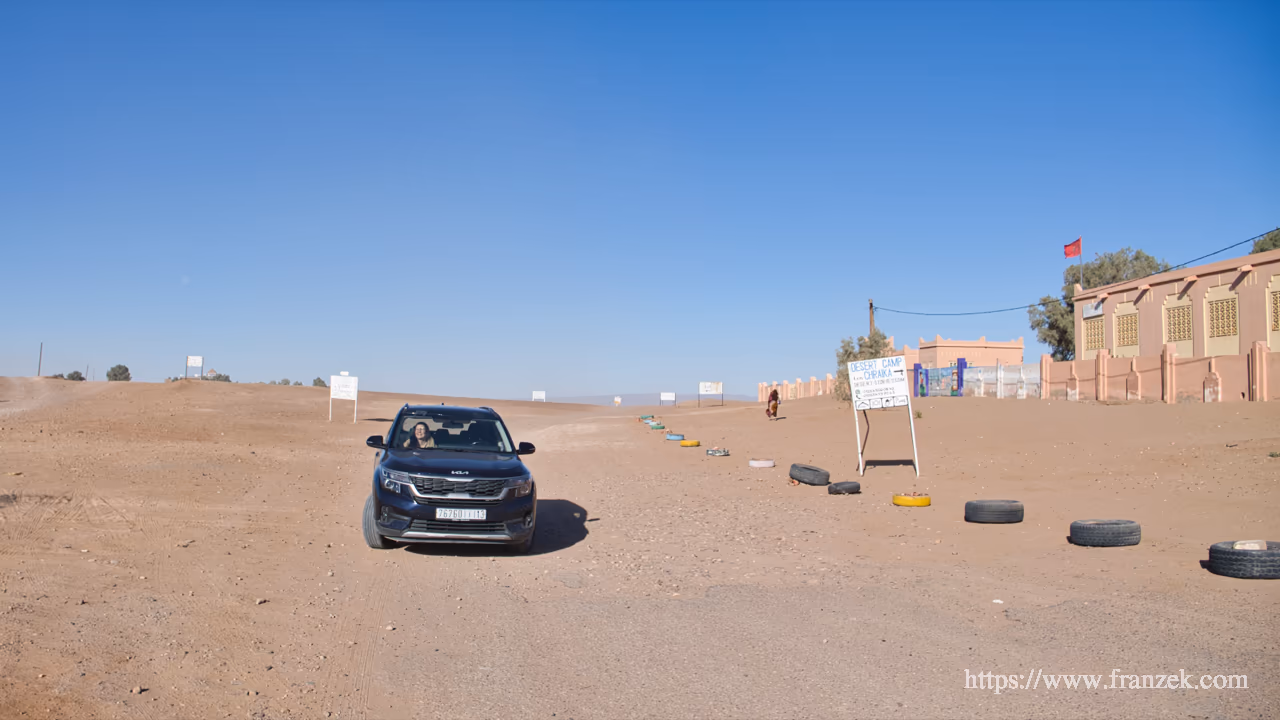 Süd-Marokko Mietwagen-Route: Kosten & Tipps (Anti-Atlas) 44 Ein dunkler SUV steht auf einem staubigen, sandigen Platz. Rechts im Hintergrund ein lachsfarbenes Gebäude mit roter Flagge, davor Hinweisschilder zu Wüstencamps und eine Begrenzung aus im Boden eingelassenen, bunt bemalten Autoreifen unter strahlend blauem Himmel.
