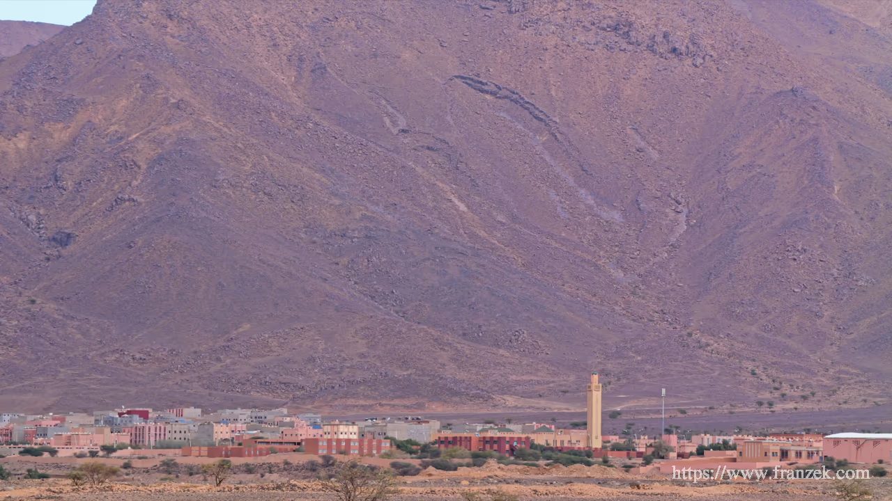 Süd-Marokko Mietwagen-Route: Kosten & Tipps (Anti-Atlas) 37 Teleaufnahme der Ortschaft Foum Zguid. Eine dichte Ansammlung von rosa und ockerfarbenen Gebäuden, aus denen ein Minarett ragt, zieht sich horizontal durch das Bild. Der gesamte Hintergrund wird von einer riesigen, steilen und dunkel-violetten Bergflanke eingenommen, die die Siedlung winzig erscheinen lässt.