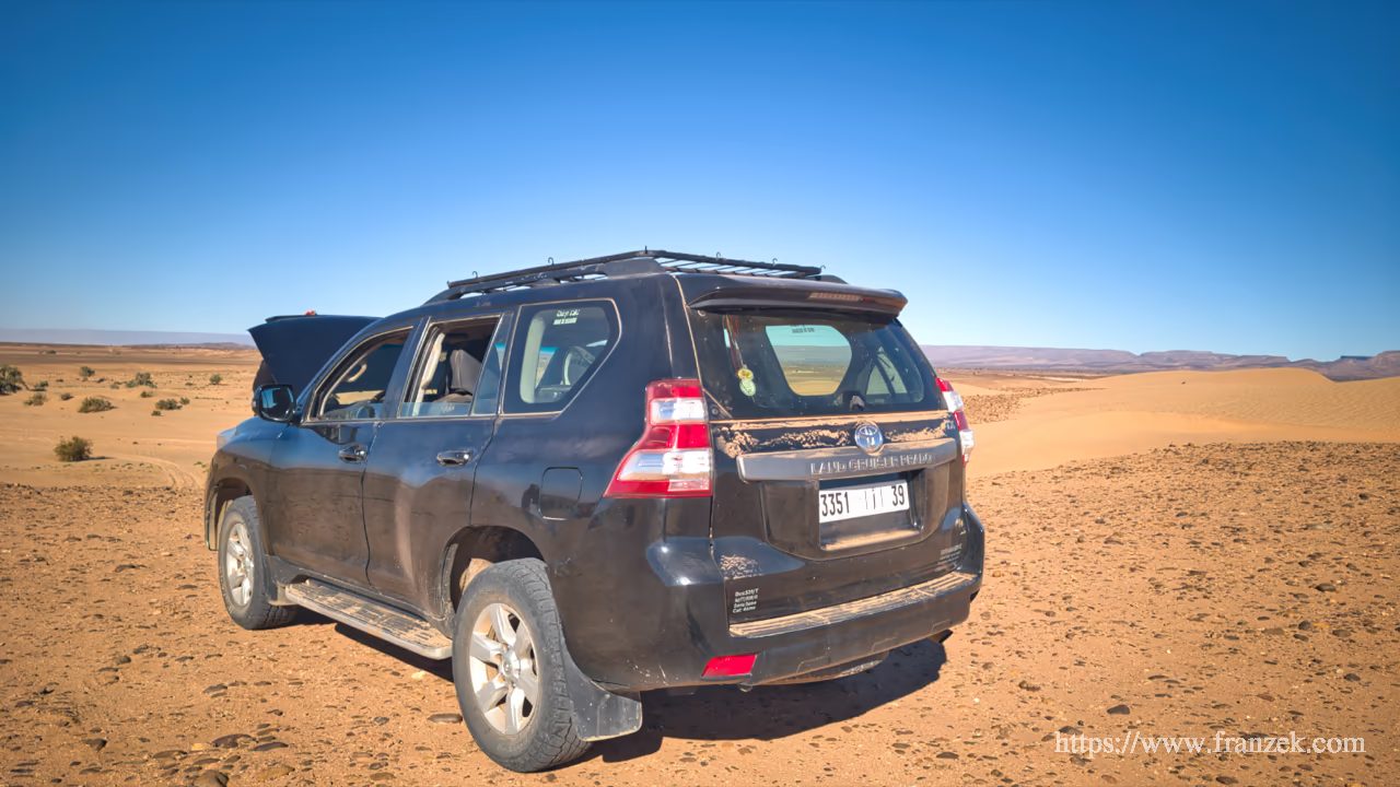 Süd-Marokko Mietwagen-Route: Kosten & Tipps (Anti-Atlas) 45 Ein schwarzer Toyota Land Cruiser parkt in einer flachen, sandigen Wüstenlandschaft unter strahlend blauem Himmel.
