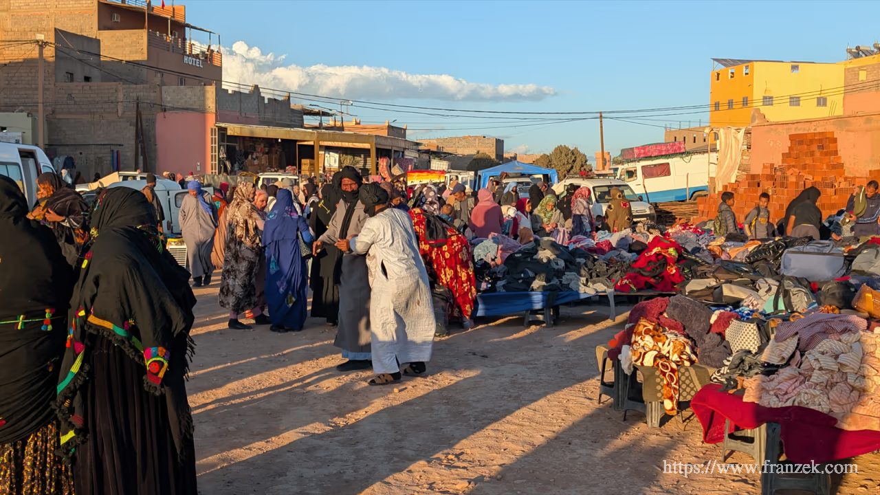 Süd-Marokko Mietwagen-Route: Kosten & Tipps (Anti-Atlas) 43 Ein belebter Marktplatz im Freien unter blauem Himmel. Menschen in traditionellen Djellabas bewegen sich zwischen improvisierten Verkaufsständen im staubigen Gegenlicht.