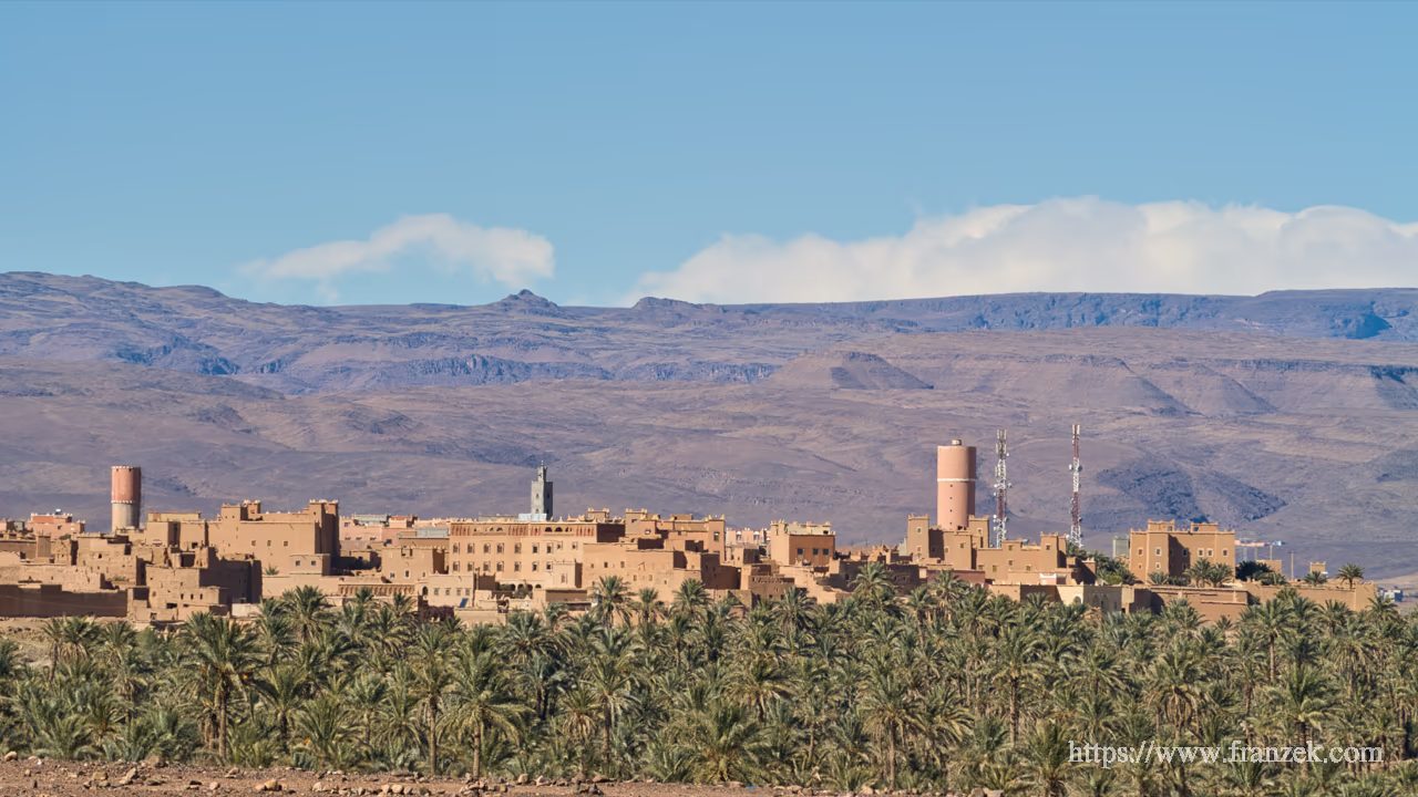 Süd-Marokko Mietwagen-Route: Kosten & Tipps (Anti-Atlas) 50 Panorama-Ansicht des Dorfes N’Kob. Zahlreiche traditionelle Kasbah-Türme ragen aus einem dichten grünen Palmenhain hervor. Im Hintergrund erstreckt sich das massive, flache Felsplateau des Jbel-Saghro-Gebirges unter blauem Himmel.