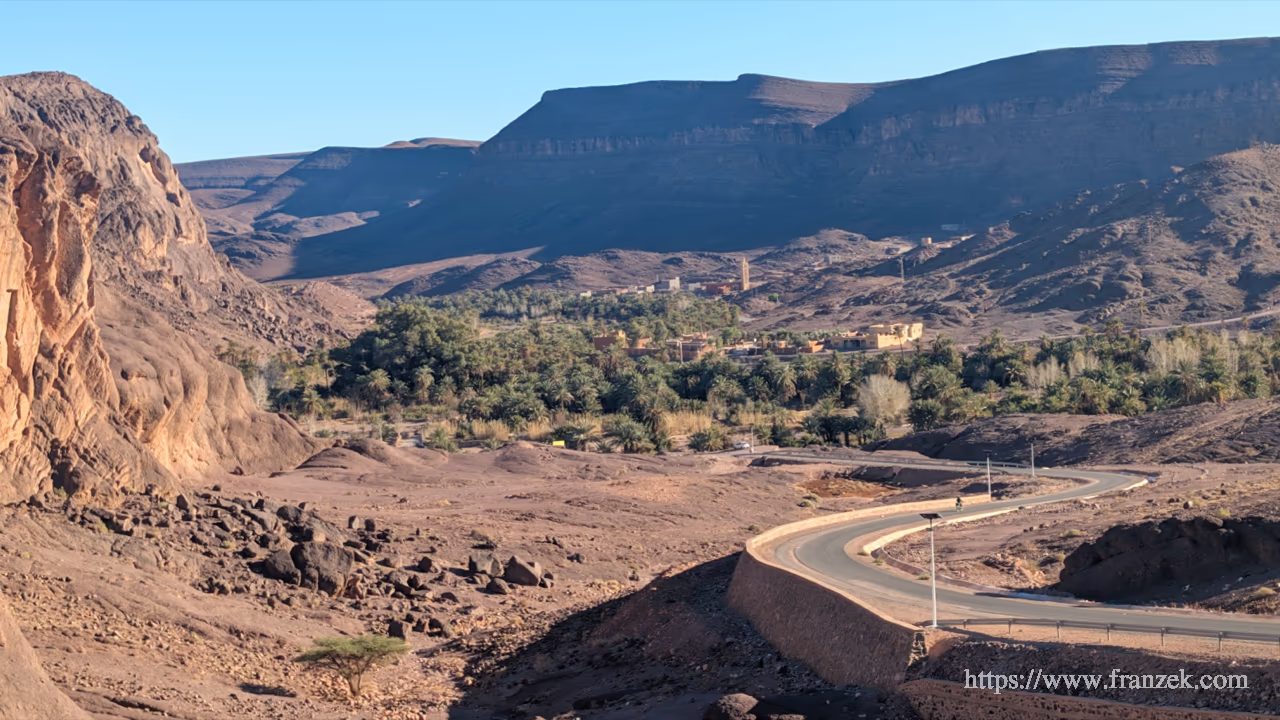 Süd-Marokko Mietwagen-Route: Kosten & Tipps (Anti-Atlas) 63 Blick über den Oued Fint, in dessen ruhigem Wasser sich die umliegenden dunklen Felswände spiegeln. Am linken Ufer parken zwei Expeditionsmobile unter einer Gruppe von Palmen und hohen, blattlosen Bäumen. Im Vordergrund ragen rötliche Steine aus dem flachen Wasser.
