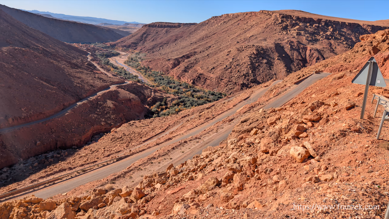 Süd-Marokko Mietwagen-Route: Kosten & Tipps (Anti-Atlas) 65 Blick von einer Anhöhe in das Ounila-Tal. Eine schmale, graue Asphaltstraße windet sich in Serpentinen durch eine tiefe Schlucht aus leuchtend rotem Fels. Am Talboden zieht sich ein schmales grünes Band aus Vegetation entlang.