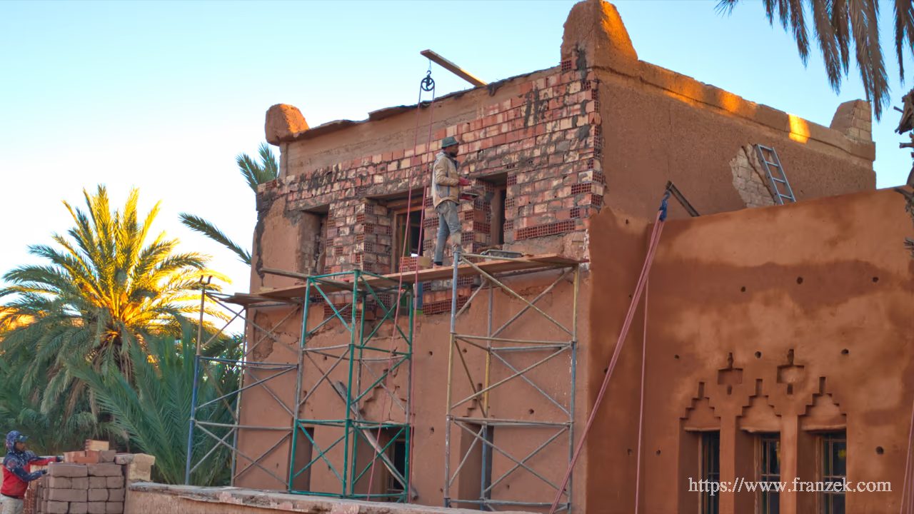 Süd-Marokko Mietwagen-Route: Kosten & Tipps (Anti-Atlas) 60 Ein Gebäude im Bau in Ouarzazate. Auf einem Holzgerüst stehen Arbeiter an einer Fassade aus rötlichem Stampflehm. Die Fensteröffnungen sind mit Ziegelsteinen verstärkt.