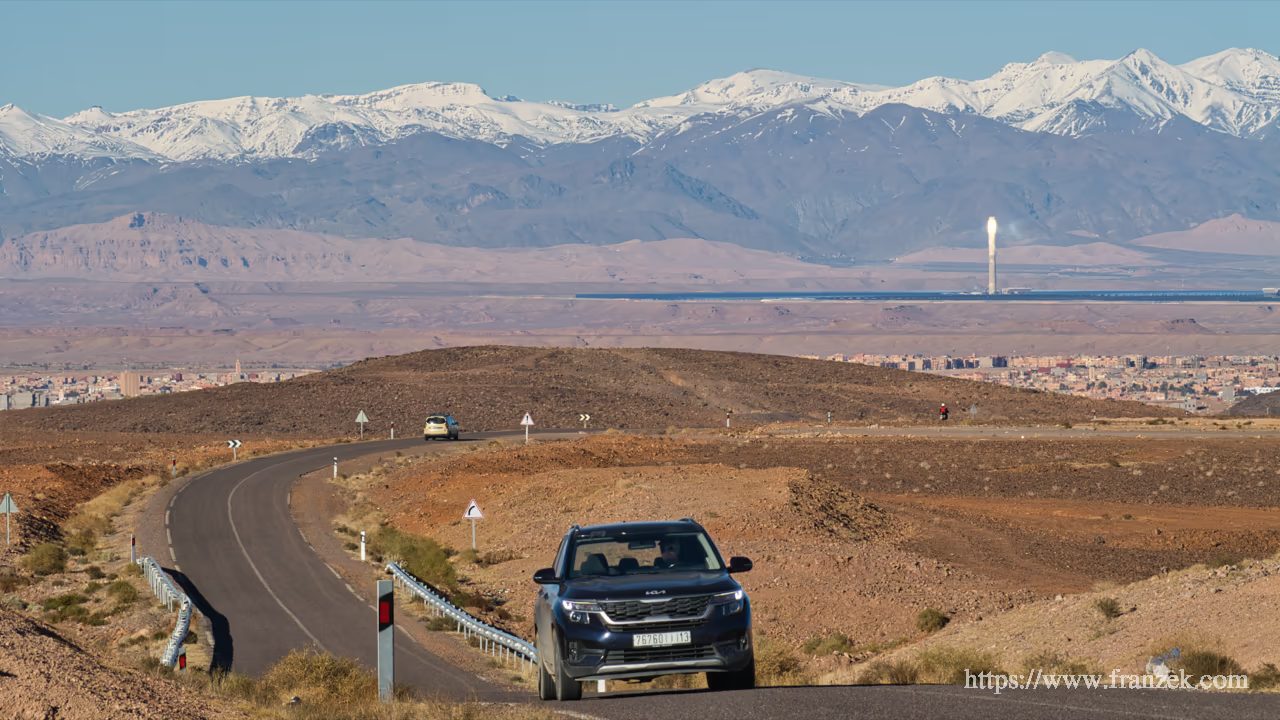 Süd-Marokko Mietwagen-Route: Kosten & Tipps (Anti-Atlas) 62 Ein dunkler SUV fährt auf einer kurvigen Asphaltstraße durch eine trockene Hügellandschaft. Im weiten Tal im Hintergrund ist die Stadt Ouarzazate und der markante, gleißend hell leuchtende Turm des Noor-Solarkraftwerks zu sehen. Den Horizont bilden die hohen, schneebedeckten Gipfel des Hohen Atlas unter klarem Himmel.