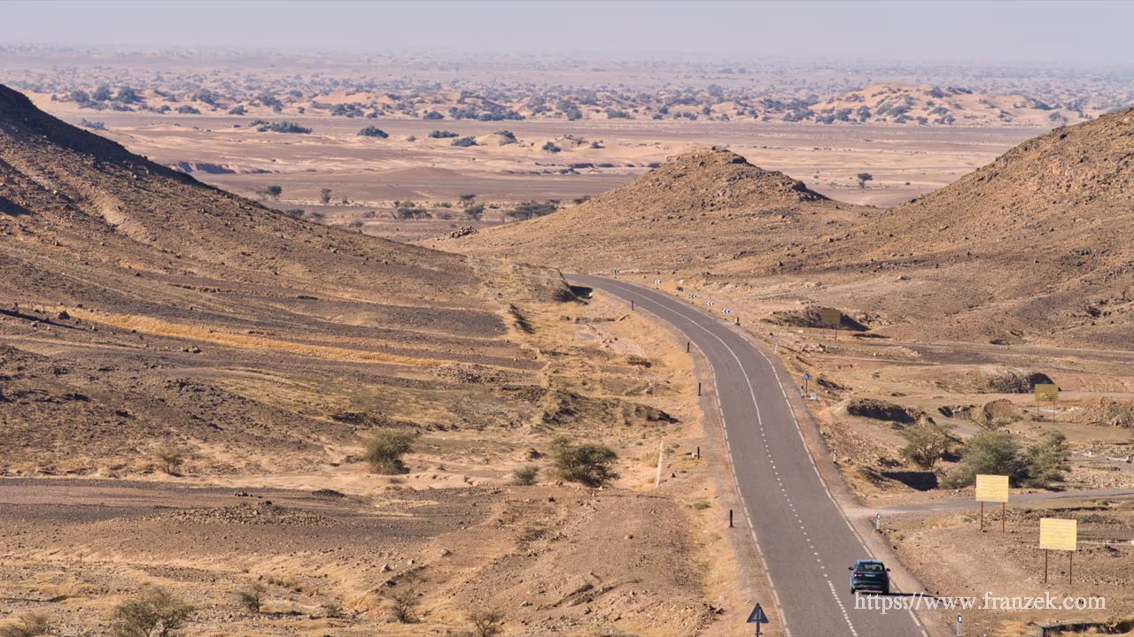 Süd-Marokko Mietwagen-Route: Kosten & Tipps (Anti-Atlas) 42 Blick von einer Anhöhe auf eine asphaltierte Straße, die sich durch eine karge, hügelige Steinwüste schlängelt. Ein dunkles Auto fährt in Richtung einer weiten Ebene mit Sanddünen im Hintergrund. Am rechten Bildrand stehen mehrere gelbe Hinweisschilder.