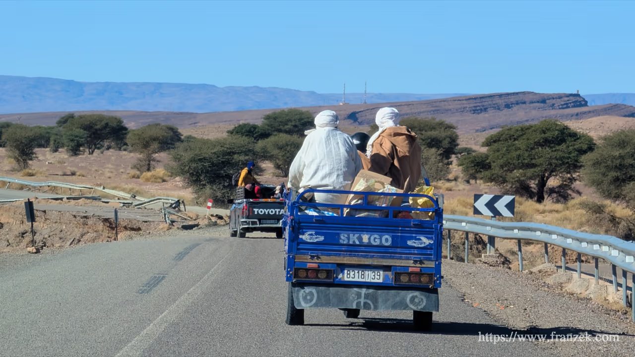 Süd-Marokko Mietwagen-Route: Kosten & Tipps (Anti-Atlas) 49 Ein blauer Pickup-Laster mit mehreren Personen auf der Ladefläche fährt auf einer asphaltierten Straße durch eine trockene Landschaft Richtung N’Kob.