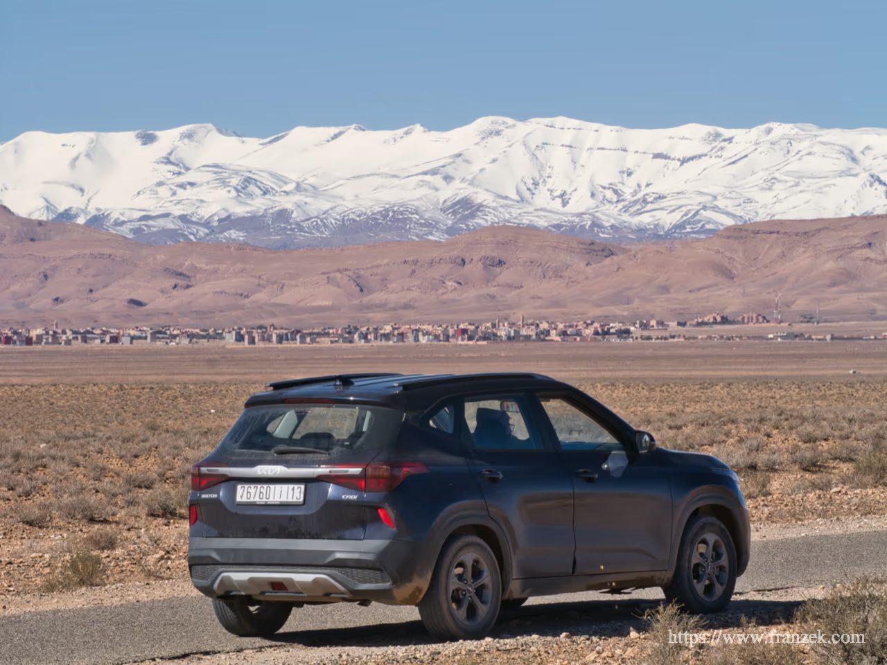 Süd-Marokko Mietwagen-Route: Kosten & Tipps (Anti-Atlas) 58 Ein dunkler SUV parkt am Rand einer asphaltierten Straße in einer weiten, trockenen Ebene. Im Hintergrund erstreckt sich die Siedlung Boulmane Dades am Fuße des massiven, tief verschneiten Hohen Atlas unter einem klaren blauen Himmel.