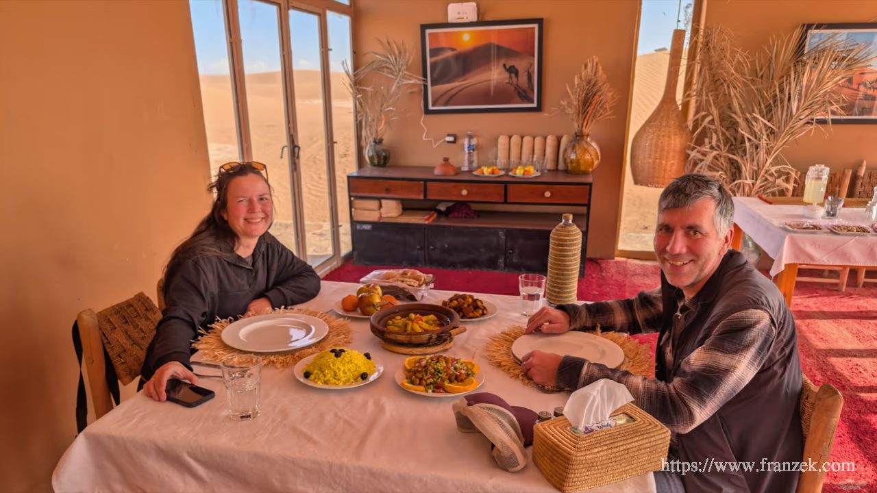Süd-Marokko Mietwagen-Route: Kosten & Tipps (Anti-Atlas) 46 Blick auf einen gedeckten Tisch mit mehreren traditionellen Tajine-Gefäßen und Beilagen in einem hellen Raum. Zwei Personen sitzen am Tisch, im Hintergrund ist ein einfaches Interieur erkennbar.