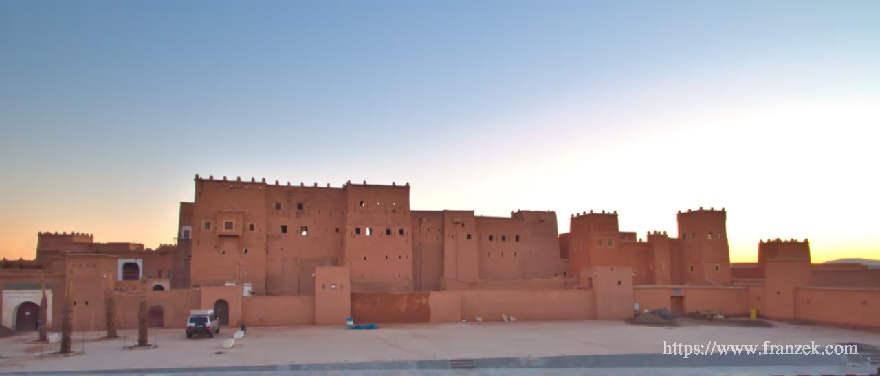 Süd-Marokko Mietwagen-Route: Kosten & Tipps (Anti-Atlas) 59 Die massive Front der Kasbah Taourirt in Ouarzazate bei einsetzender Dämmerung. Die hohen, fensterarmen Lehmwände und Wehrtürme der ehemaligen Glaoui-Residenz sind in rötliches Licht getaucht.