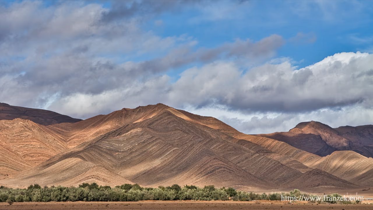 Süd-Marokko Mietwagen-Route: Kosten & Tipps (Anti-Atlas) 30 Blick auf eine karge Gebirgskette mit markanten, diagonal verlaufenden Gesteinsschichten, die dreieckige Faltungen bilden. Am Fuß der braun-ockerfarbenen Berge verläuft ein dichter grüner Vegetationsstreifen aus Bäumen und Büschen. Darüber ein blauer Himmel mit lockerer Bewölkung.