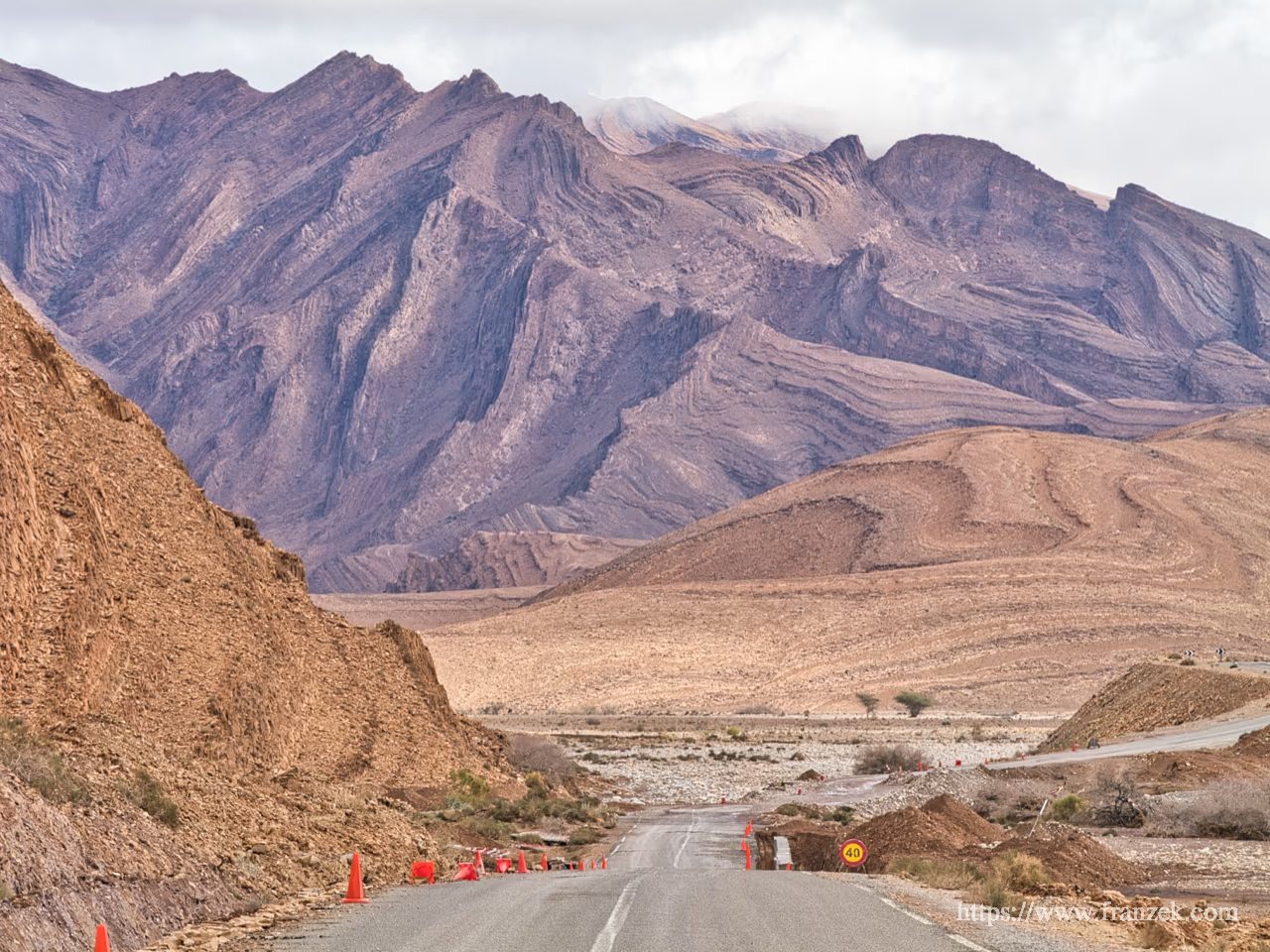 Süd-Marokko Mietwagen-Route: Kosten & Tipps (Anti-Atlas) 29 Eine asphaltierte Landstraße führt geradeaus durch eine karge, steinige Ebene auf eine massive Gebirgskette zu. Im Vordergrund begrenzen orangefarbene Leitkegel die Fahrbahn. Die Berge im Hintergrund dominieren das Bild mit spektakulären, violett-braunen Gesteinsschichten, die extrem stark gefaltet und verbogen sind und wie riesige, versteinerte Wellen aussehen. Der Himmel ist bewölkt.