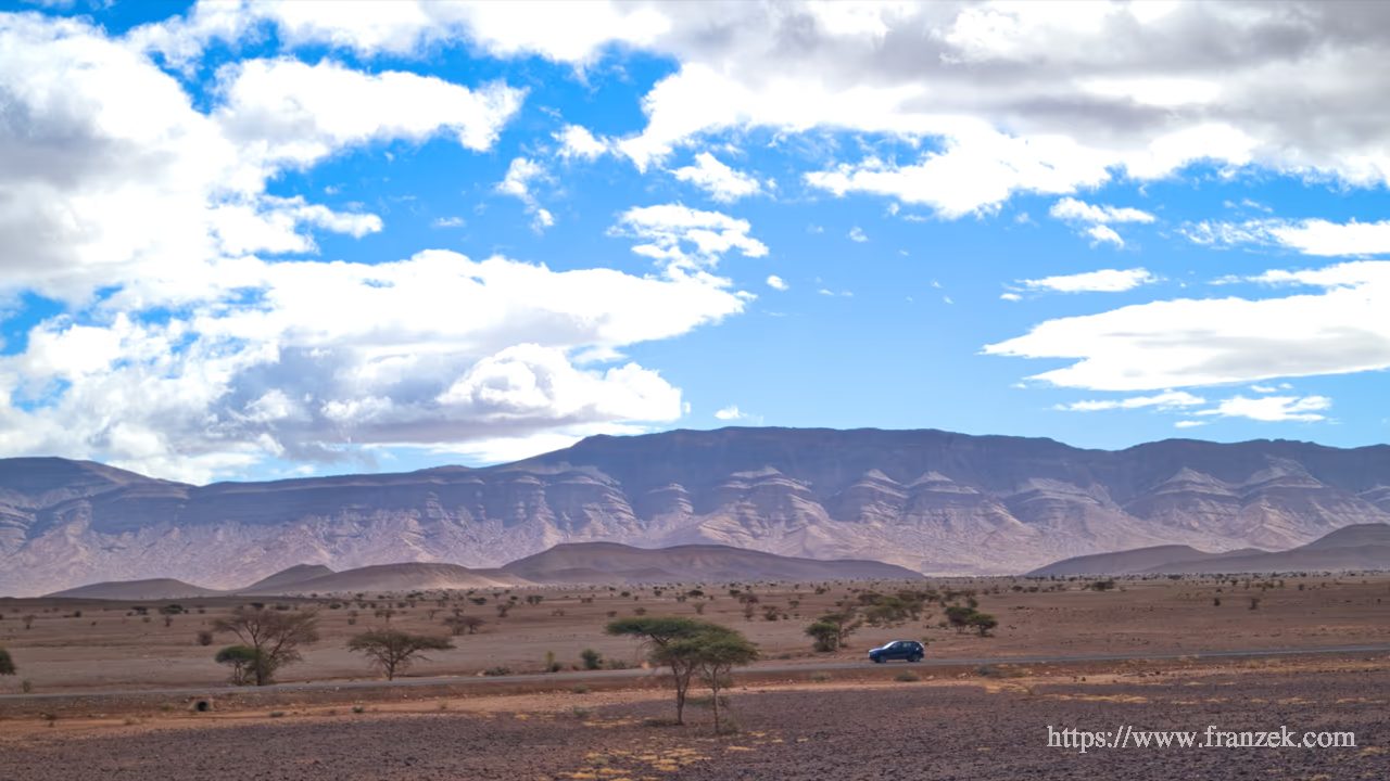 Süd-Marokko Mietwagen-Route: Kosten & Tipps (Anti-Atlas) 35 Weitwinkelaufnahme der Route P1800 in einer kargen Ebene. Ein blauer Geländewagen fährt auf der schmalen Asphaltstraße, umgeben von vereinzelten Akazienbäumen. Im Hintergrund erstreckt sich eine lange, massive Gebirgskette unter einem weiten Himmel mit weißen Wolken.