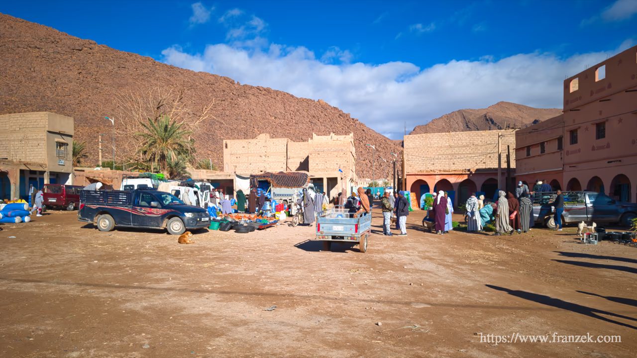 Süd-Marokko Mietwagen-Route: Kosten & Tipps (Anti-Atlas) 36 Belebter, staubiger Marktplatz in Tissint vor einer massiven, rötlichen Felswand. Auf dem Platz stehen mehrere Pickups und Transporter, dazwischen Männer in traditioneller Kleidung. Im Vordergrund ruht ein Hund im Schatten eines Fahrzeugs.