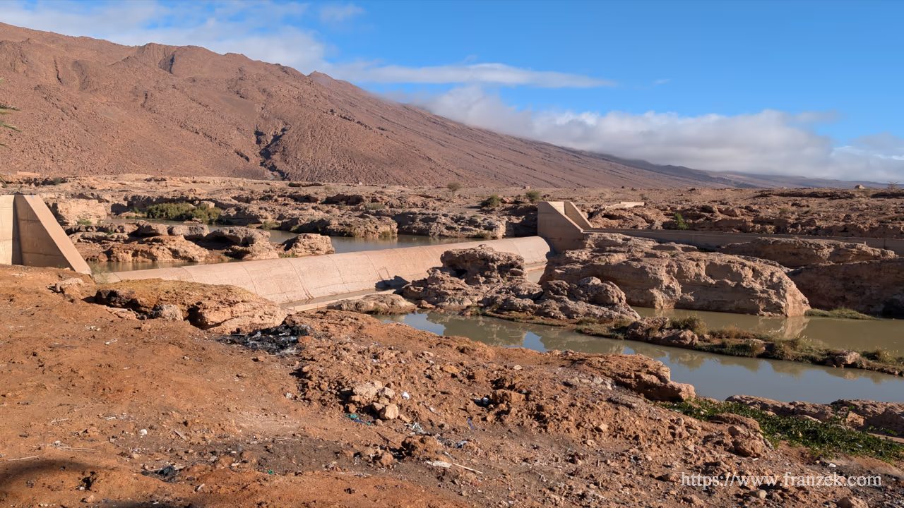 Süd-Marokko Mietwagen-Route: Kosten & Tipps (Anti-Atlas) 34 Eine betonierte Staustufe (Wehr) zieht sich quer durch ein felsiges Flussbett. Das Wasser staut sich vor der Barriere und fließt seitlich weiter. Die Umgebung ist karg und steinig, im Hintergrund rötliche Berghänge.