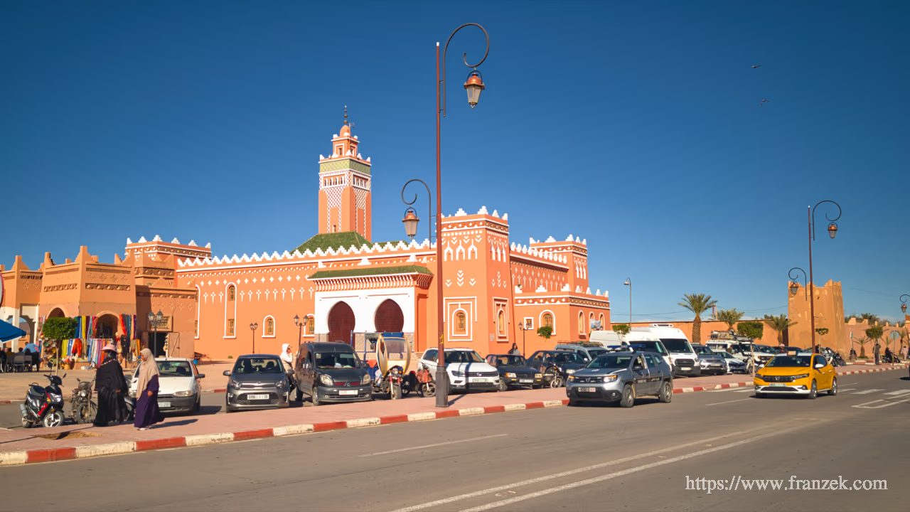 Marokko im Ramadan mit dem Wohnmobil: Was du wissen musst 4 Stadtansicht im Zentrum von Zagora. Ein großes, lachsfarbenes Gebäude mit Zinnen und Turm (im Stil einer Kasbah) dominiert den Platz. Davor ein asphaltierter Parkplatz mit modernen Autos und Straßenlaternen.