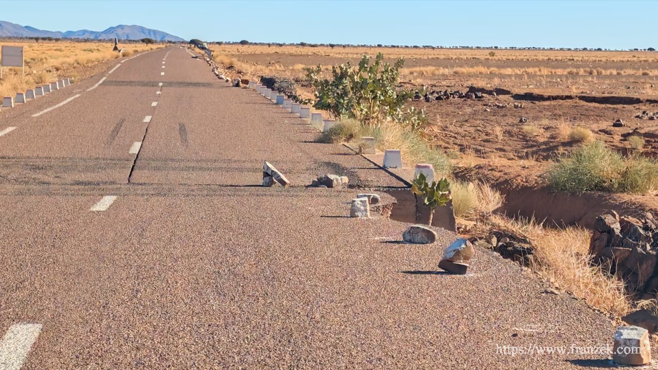 Süd-Marokko Mietwagen-Route: Kosten & Tipps (Anti-Atlas) 39 Nahaufnahme einer ausgefransten, abgebrochenen Asphaltkante einer Landstraße. Mehrere grobe Felsbrocken wurden als improvisierte Warnmarkierung direkt auf den Straßenbelag gelegt.
